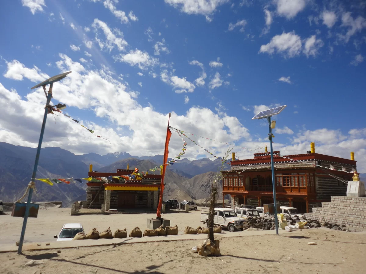 Nako Monastery Spiti Valley during partly cloudy skies, featuring red buildings prayer flags solar panels vehicles barren mountains courtyard, perfect spiritual experience Himachal Pradesh tour package.