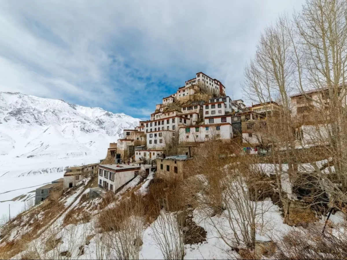 Multi-tiered Nako Monastery at Kinnaur during winter partly cloudy, featuring white washed buildings, snowy mountains, barren trees, perfect spiritual Himachal Pradesh tour package.