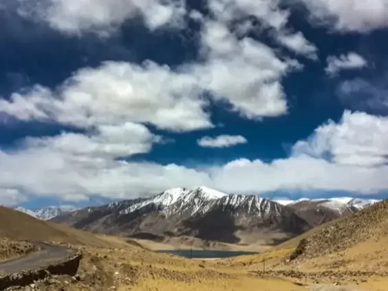 Namashang La Pass Ladakh during partly cloudy day, featuring snow-capped peaks turquoise lake valley, winding road barren hills blue sky, perfect scenic adventure Ladakh tour package.