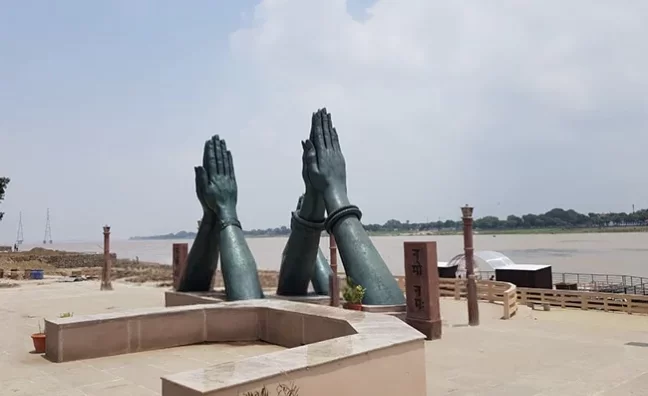 Namaste Ghat Varanasi daytime, giant green bronze prayer hands bangles Ganga river pillars plants sandy bank, perfect Uttar Pradesh tour package.