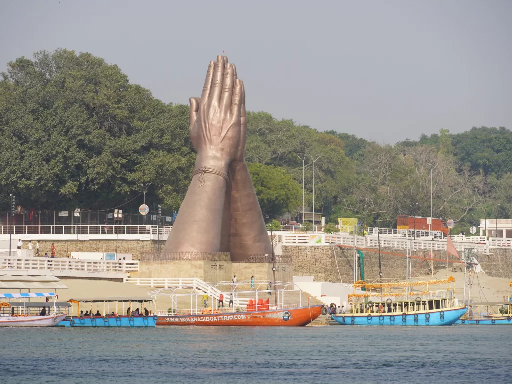 Namaste Ghat Varanasi daytime, giant bronze prayer hands bangles boats river trees ghats people, perfect Uttar Pradesh tour package.
