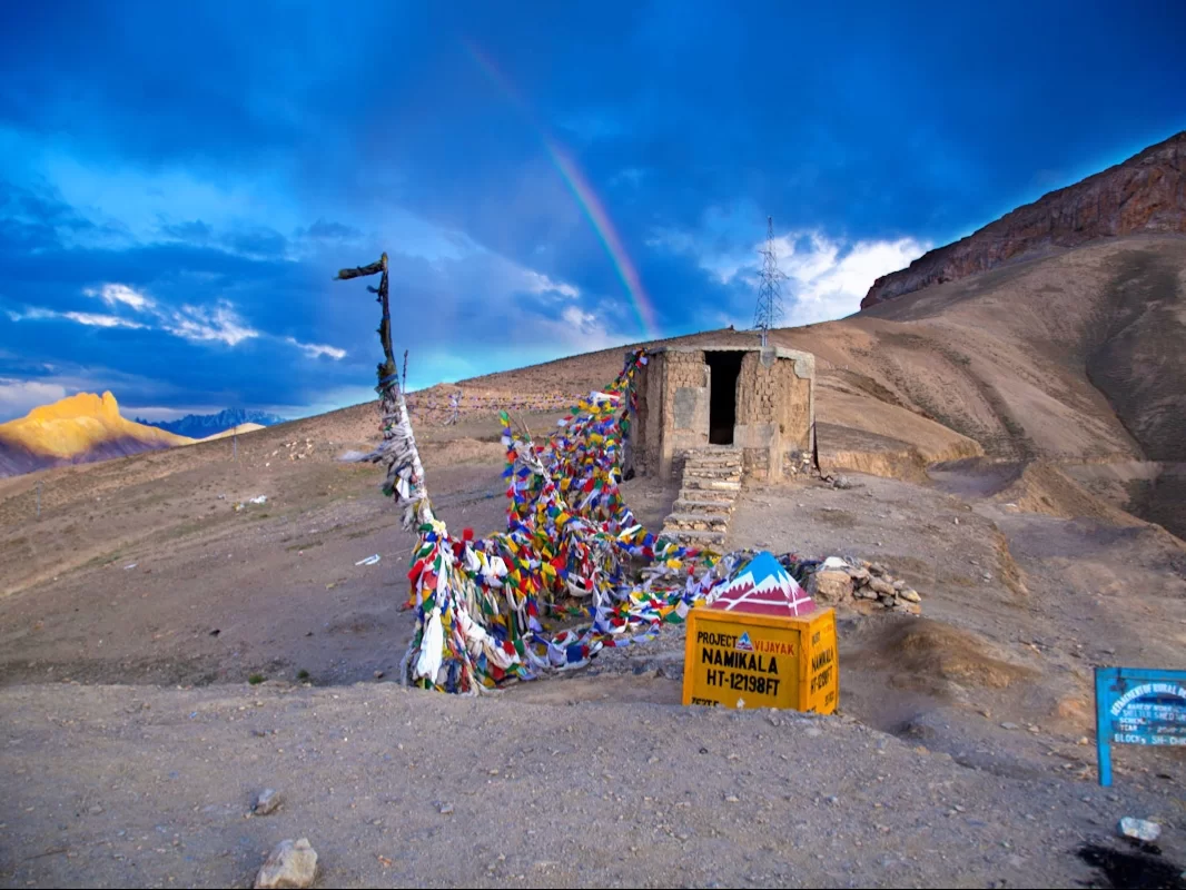 Namika La Pass summit Ladakh with Maitreya Buddha statue prayer flags stone hut BRO sign during partly cloudy day, featuring dramatic Zanskar mountains, perfect spiritual high-altitude Ladakh tour package