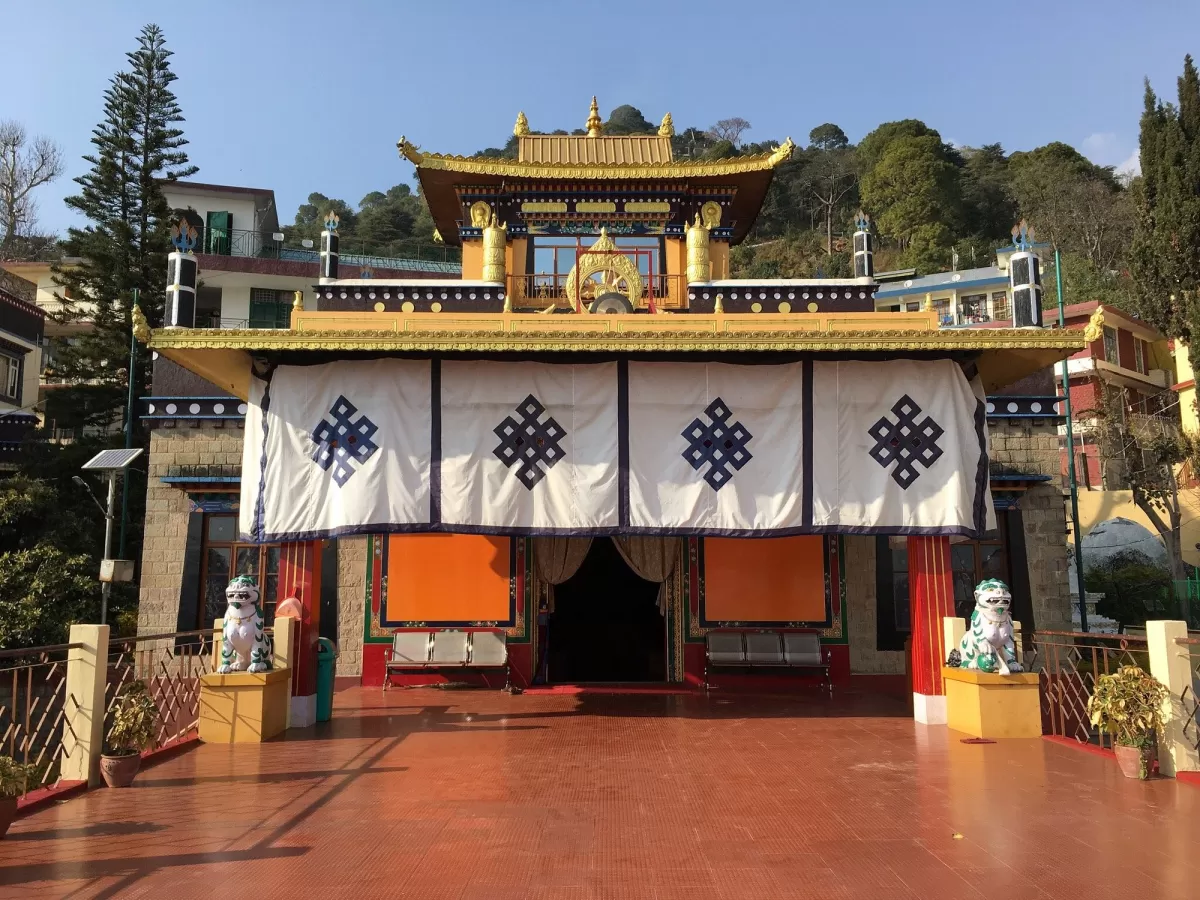 Nechung Monastery in Dharamshala featuring a traditional Tibetan-style temple with golden roof details, colorful facade, lion statues at the entrance, and hillside buildings in the background.