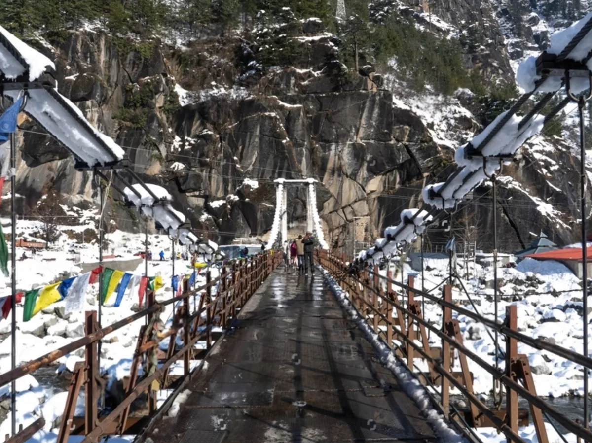Suspension bridge at Nehru Kund Manali during snowy weather, featuring prayer flags rocky cliffs and distant peaks, perfect adventure experience Himachal Pradesh tour package.