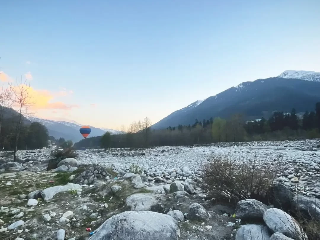 Hot air balloon over riverbed at Nehru Kund Manali during golden hour sunset, featuring rocky terrain snowy peaks and barren trees, perfect adventure experience Himachal Pradesh tour package.