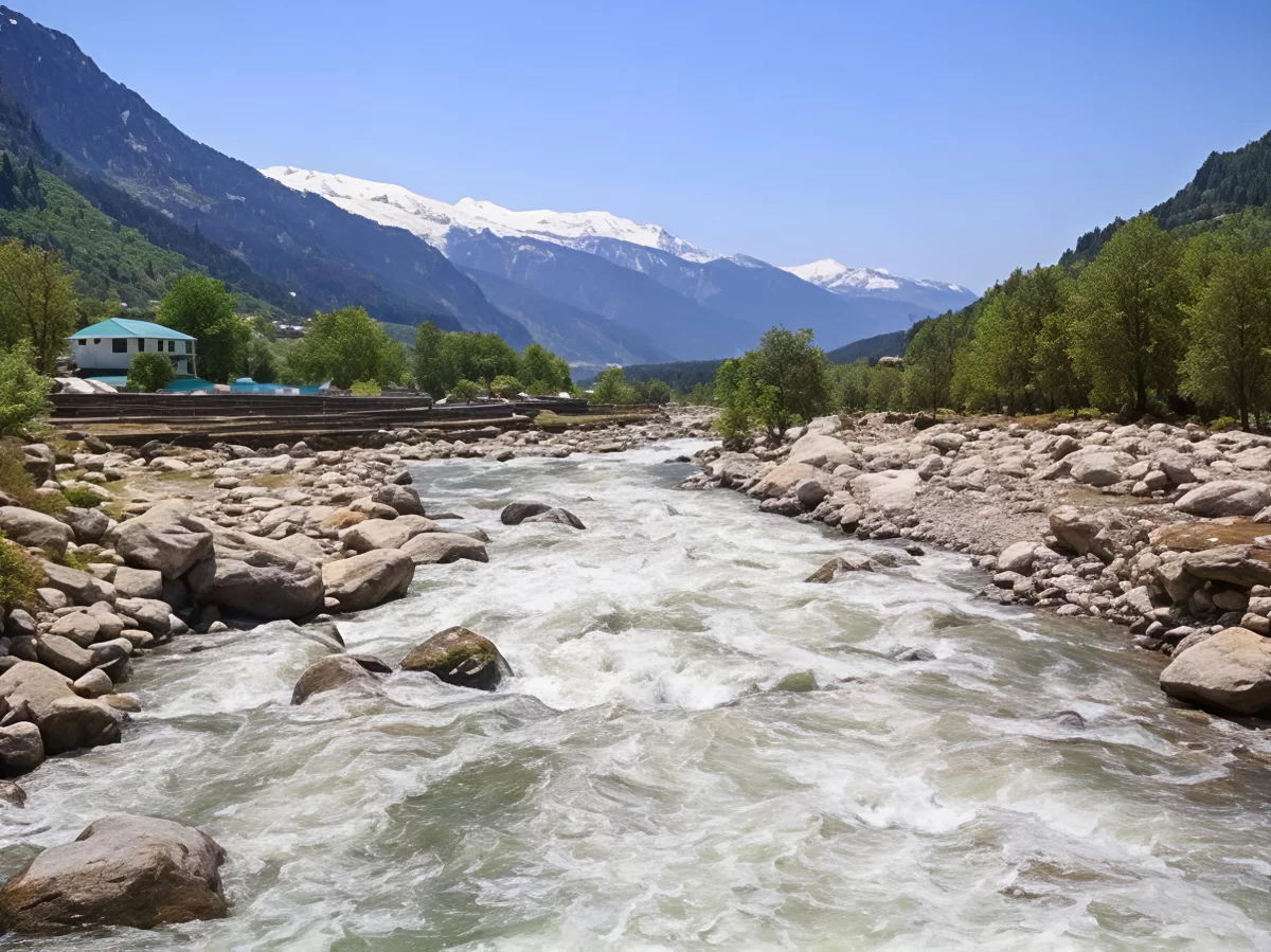 Flowing river at Nehru Kund Manali during sunny skies, featuring rocky riverbed pine trees and snow-capped peaks, perfect nature experience Himachal Pradesh tour package.