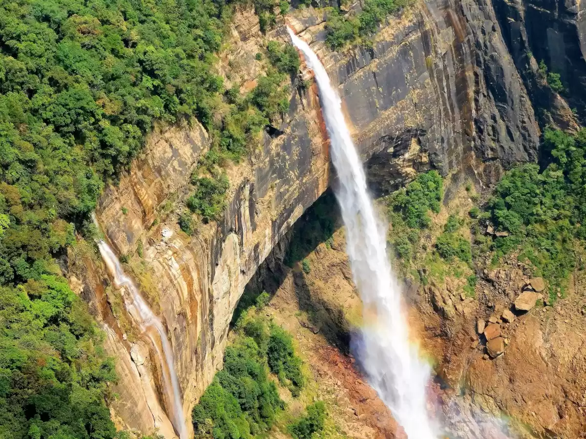 Nohkalikai Falls Cherrapunji during sunny day, featuring waterfalls cave rainbow misty spray green forested cliffs rocks, perfect scenic adventure Cherrapunji Meghalaya tour package.
