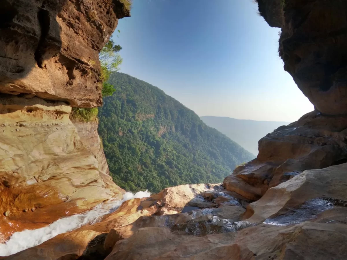 Nohkalikai Falls Cherrapunji during golden hour, featuring cave viewpoint rocky cliffs cascading waterfall lush green forested valleys blue sky, perfect scenic adventure Cherrapunji Meghalaya tour package.