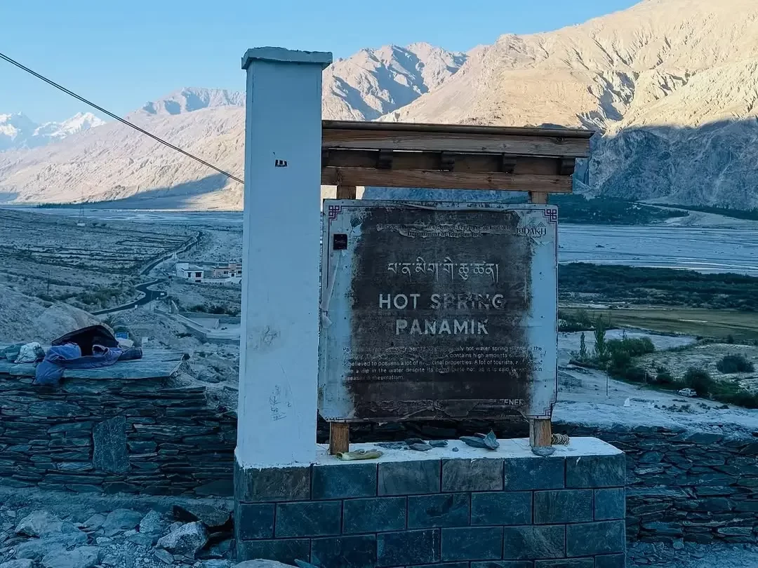 Panamik Hot Springs signboard in Nubra Valley Ladakh during golden hour sunset, featuring sulfur pool gate, snow peaks, Shyok River valley, perfect therapeutic Panamik tour package. 