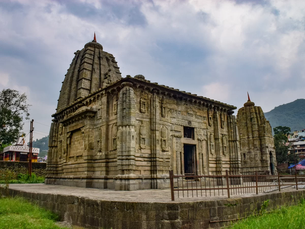 Historic Panchavaktra Temple featuring intricate stone carvings, tall shikhara towers with red flags, and ancient architectural detailing set against a cloudy sky and surrounding hills.