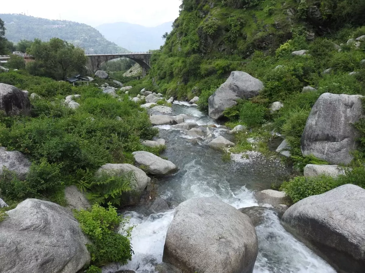 Panchpula waterfall bridge at Dalhousie during misty weather, featuring rushing stream, boulders, green hills, perfect adventure Himachal Pradesh tour package.