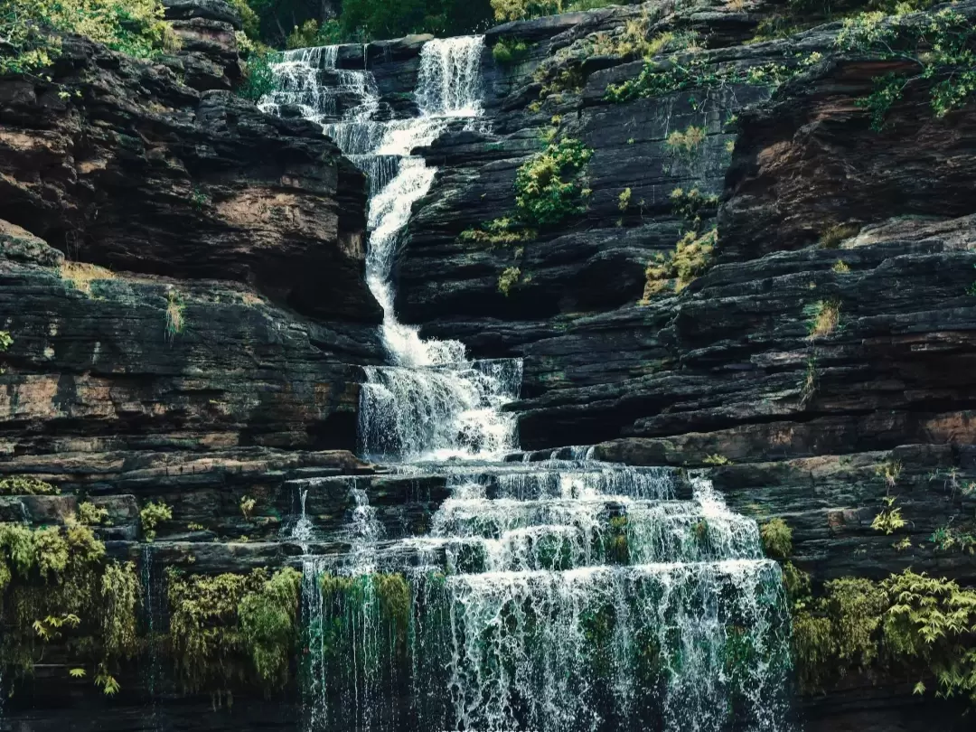 Pandav Falls in Madhya Pradesh, a multi-tiered waterfall cascading over dark layered rock formations surrounded by lush greenery, a scenic natural attraction featured in Madhya Pradesh tour packages.