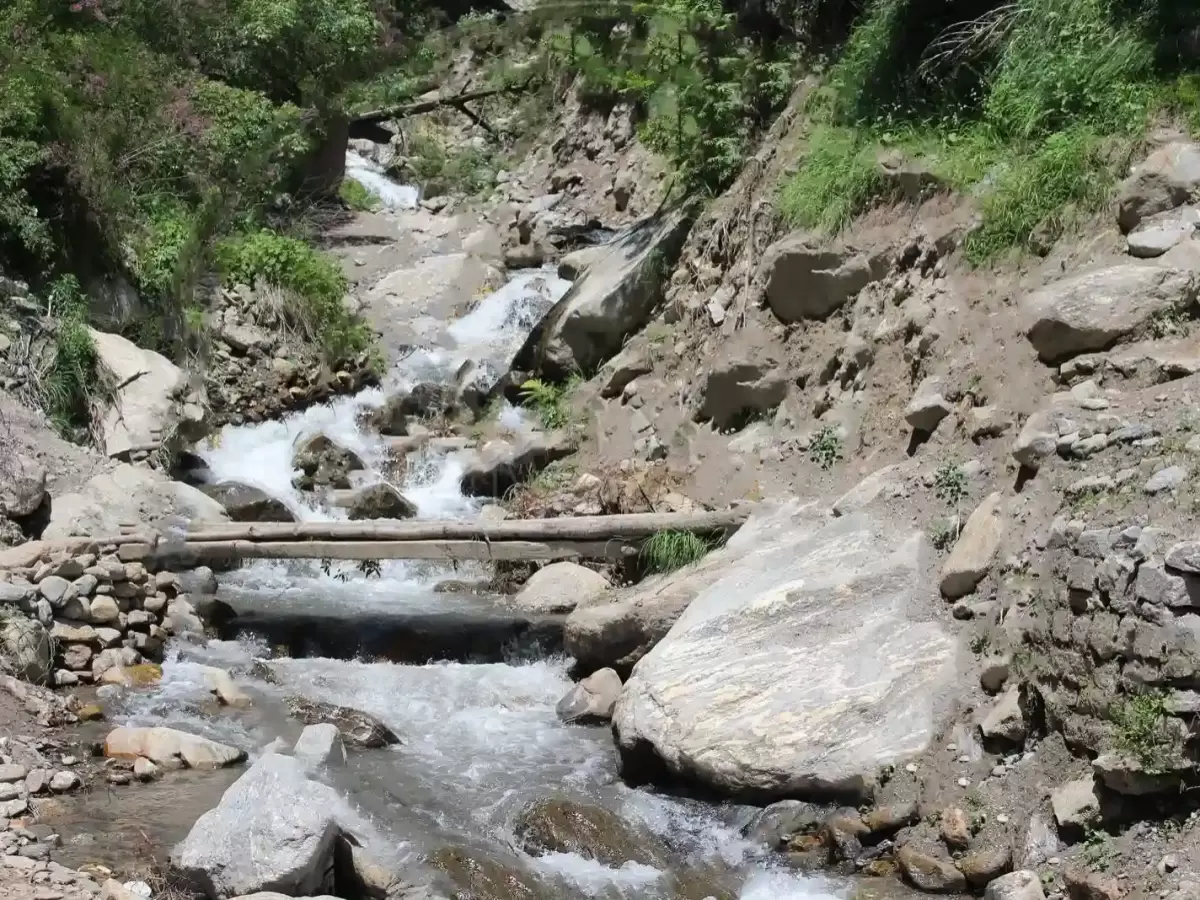 Parvati River tributary Kasol Parvati Valley Himachal Pradesh during sunny weather, featuring cascading waterfalls wooden stone bridge green forested rocky canyon backdrop, perfect cultural experience Himachal tour package.