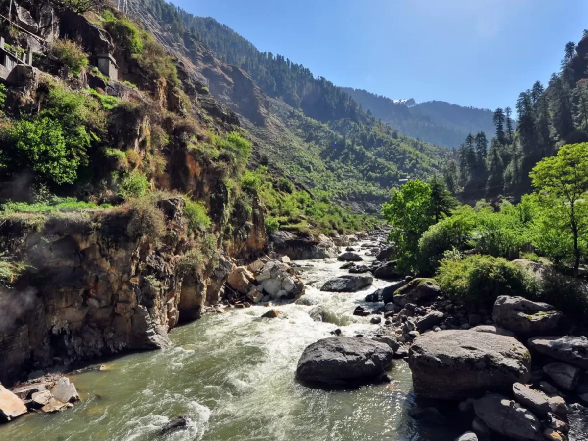 Parvati River Kasol Parvati Valley Himachal Pradesh during sunny blue skies, featuring turquoise rapids large boulders green forested rugged brown Himalayan cliffs backdrop, perfect cultural experience Himachal tour package.
