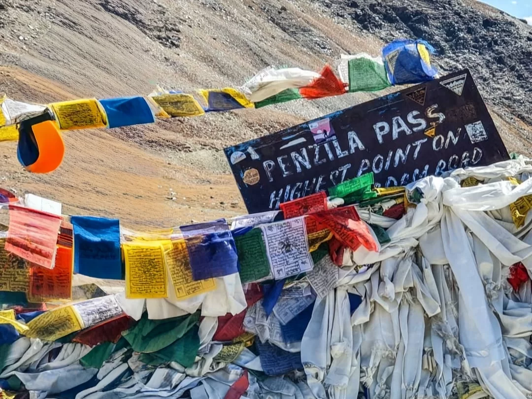 Penzila Pass Pensi La highest point signboard colorful prayer flags barren rocky slopes blue sky Kargil Ladakh Zanskar region, perfect iconic high-altitude pass adventure Ladakh tour package.