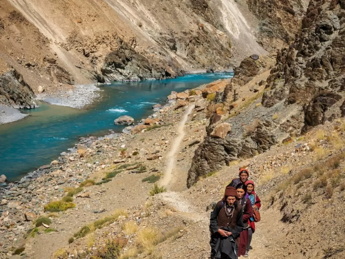 Nomads trekking Phuktal trail at Tsarap River Zanskar during clear day, featuring turquoise waters, rugged gorges, prayer flags, perfect adventure experience with Phuktal trek package