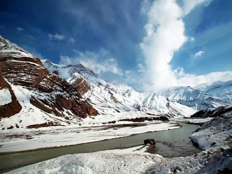 Pin Valley near Kaza Spiti winter landscape, featuring snow-covered rugged mountains turquoise river valley dramatic clouds blue skies, perfect Himachal Pradesh tour packages.