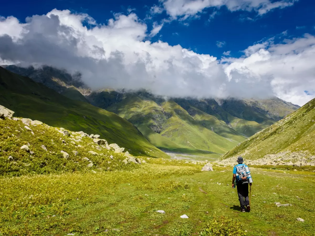 Trekking in Pin Valley National Park near Kaza during partly cloudy skies, featuring hiker green meadows rocky valley snow mountains, perfect adventure experience Himachal tour package.