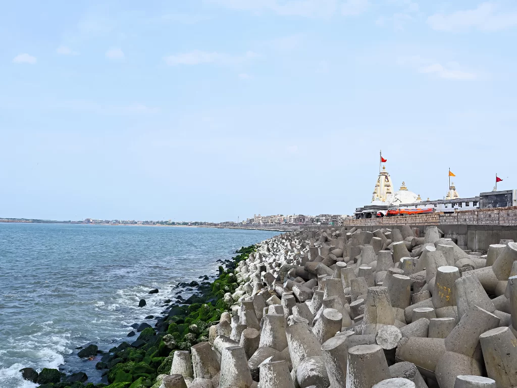 Nishkalank Mahadev Temple at Koliyak Beach Bhavnagar during clear skies, featuring white shikharas, saffron flags, sea waves and rocky shoreline, perfect spiritual experience with Gujarat tour packages.