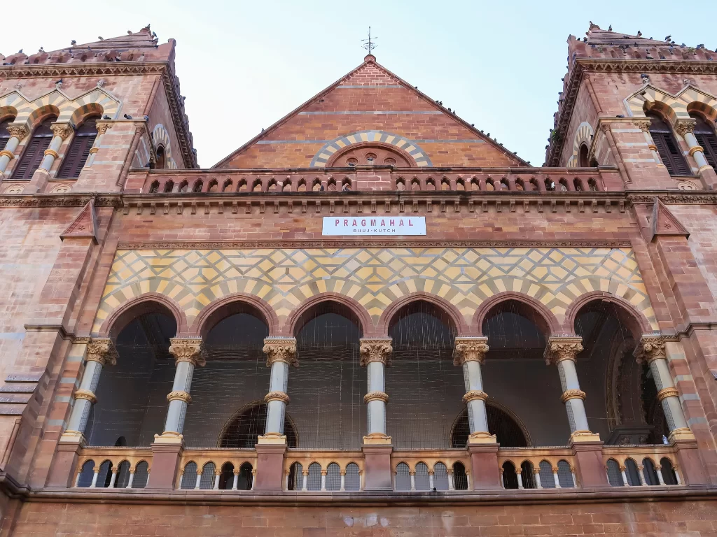 Prag Mahal main facade in Bhuj, Kutch during clear day, featuring Italian Gothic architecture with red sandstone arches, Corinthian columns, intricate jali work and PRAG MAHAL signboard, perfect heritage landmark with Gujarat tour packages.​