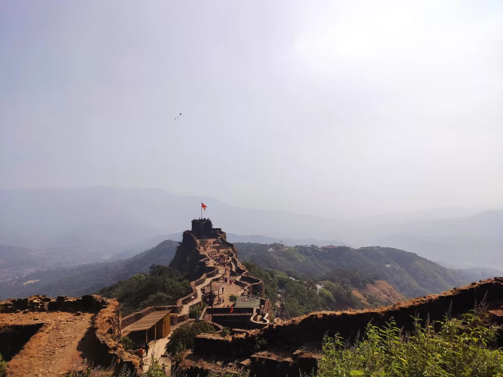 Pratapgad Fort Mahabaleshwar elevated trails, featuring winding ramparts red saffron flag atop peak misty valleys green hills hazy skies, perfect Maharashtra tour packages.