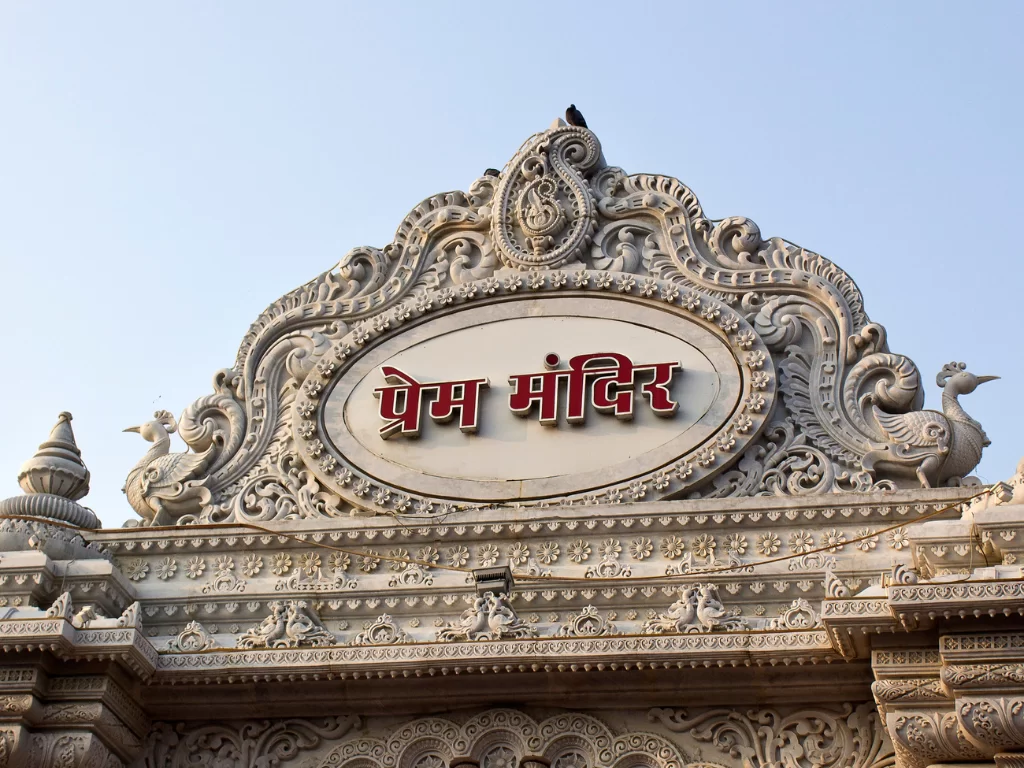 Prem Mandir features ornate white marble entrance signage in Hindi and Devanagari script, flanked by intricate carvings of peacocks and floral motifs under a clear sky, ideal for Mathura Vrindavan heritage tours