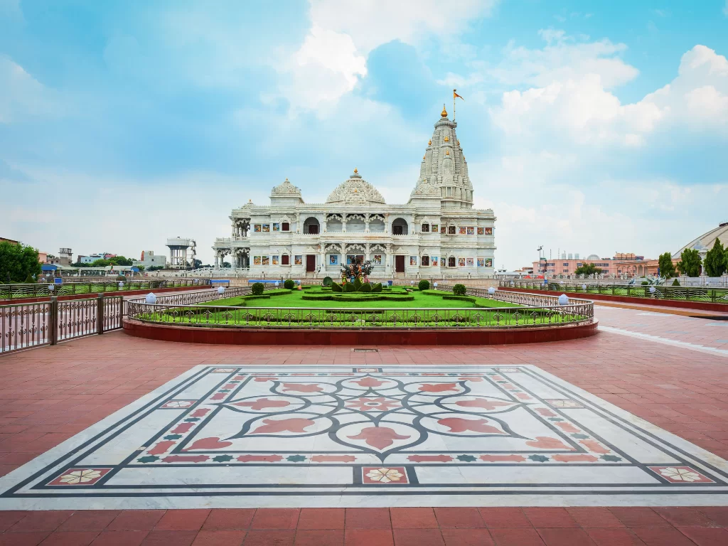 Prem Mandir's grand white marble facade under blue skies with clouds, showcasing ornate arches, golden spires, and landscaped gardens with intricate terracotta flooring, ideal for spiritual heritage tours in Vrindavan.