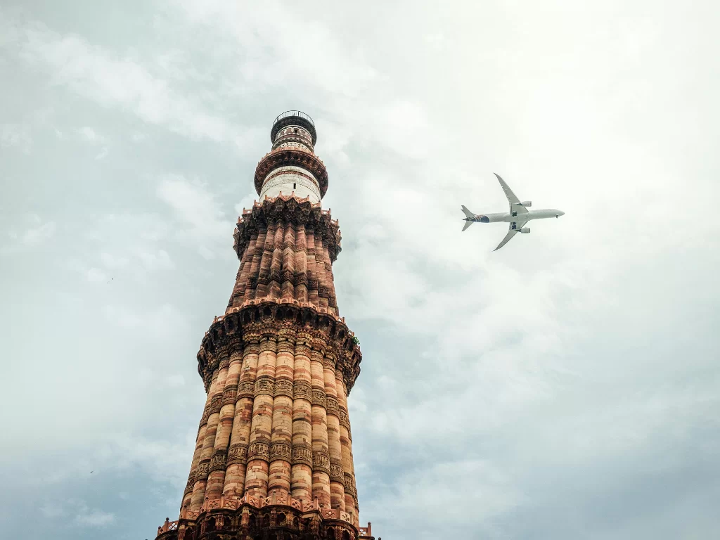 Qutub Minar close-up against cloudy sky with airplane flying overhead, showcasing Delhi’s iconic UNESCO World Heritage sandstone minaret and its blend of ancient architecture with modern air travel.
