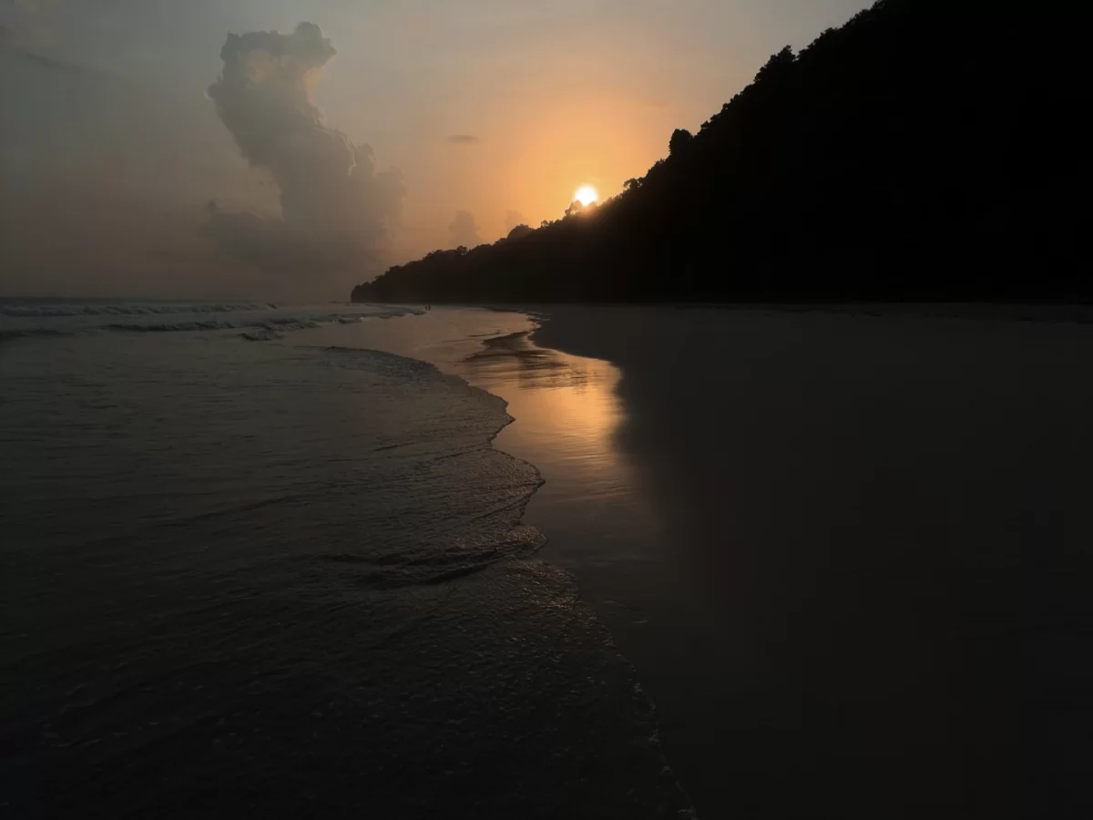 Radhanagar Beach No.7 Havelock Island Andaman sunset silhouette with setting sun behind misty clouds and forested hills, featuring reflective waves on white sand beach, tropical paradise tour package. 