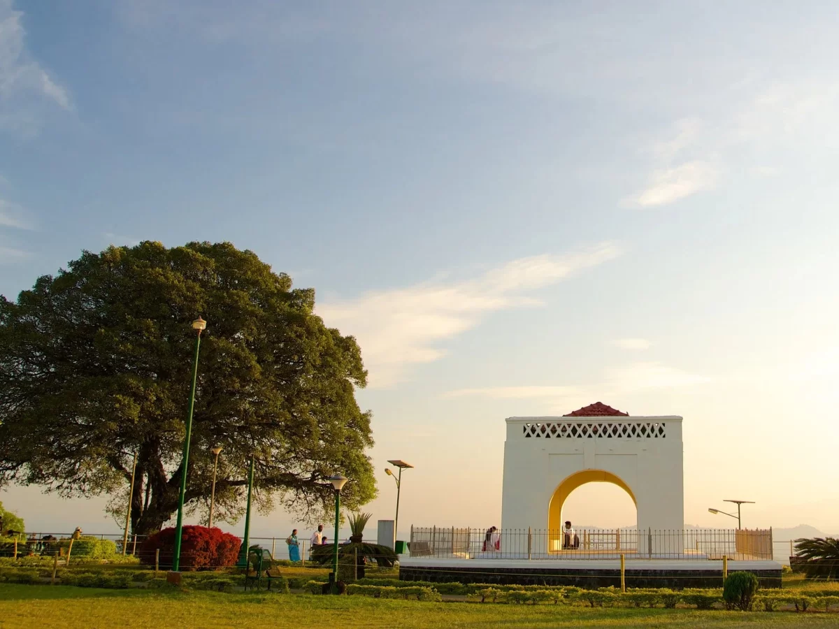 Raja Seat Garden at Madikeri Coorg during sunset, featuring iconic white arch monument, banyan tree, lamps, gardens, tourists, perfect romantic experience Karnataka tour packages.