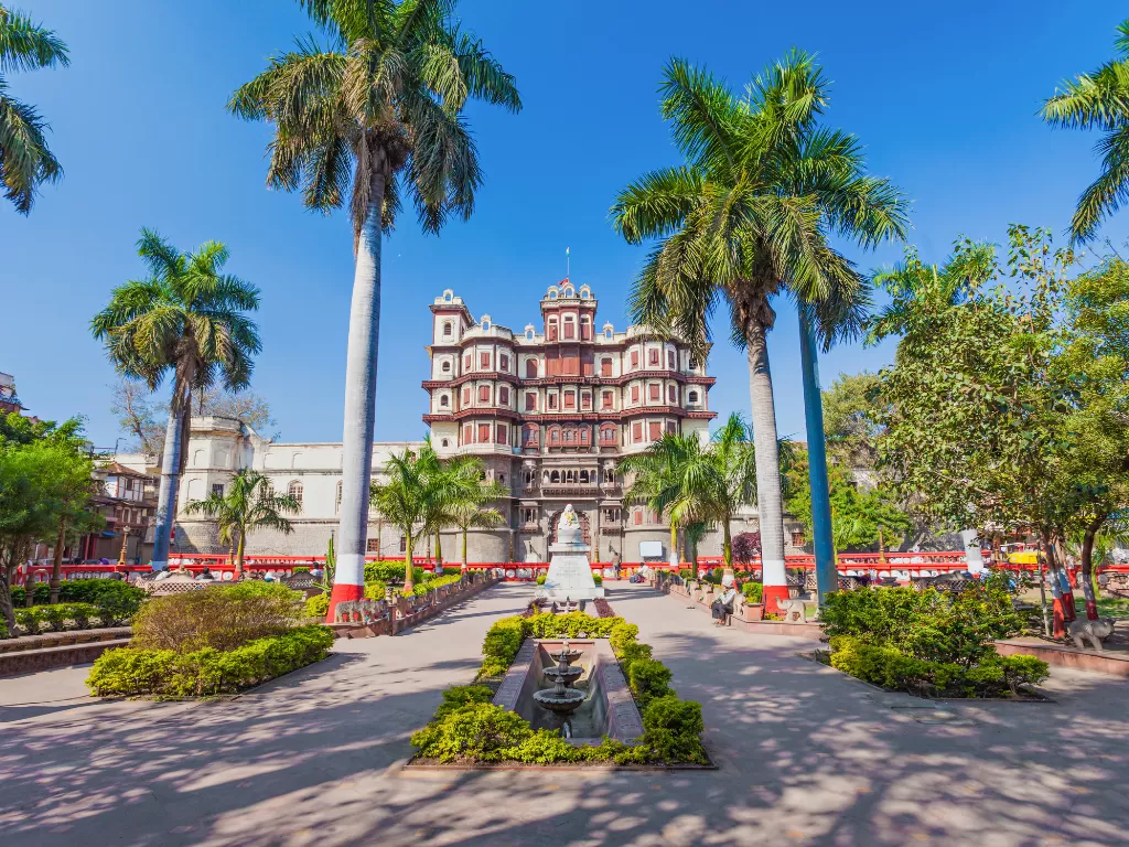 Rajwada Palace at Indore during clear daylight, featuring red-white palace tall palms garden fountain, perfect cultural experience Madhya Pradesh tour package.