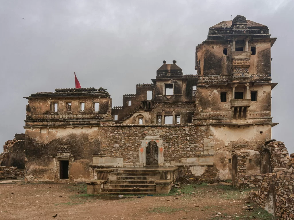 Frontal view of Rana Kumbha Palace ruins at Chittorgarh Fort in Rajasthan with multi-tiered domes, red flag and arched entrance under overcast sky, perfect Rajasthan tour package.