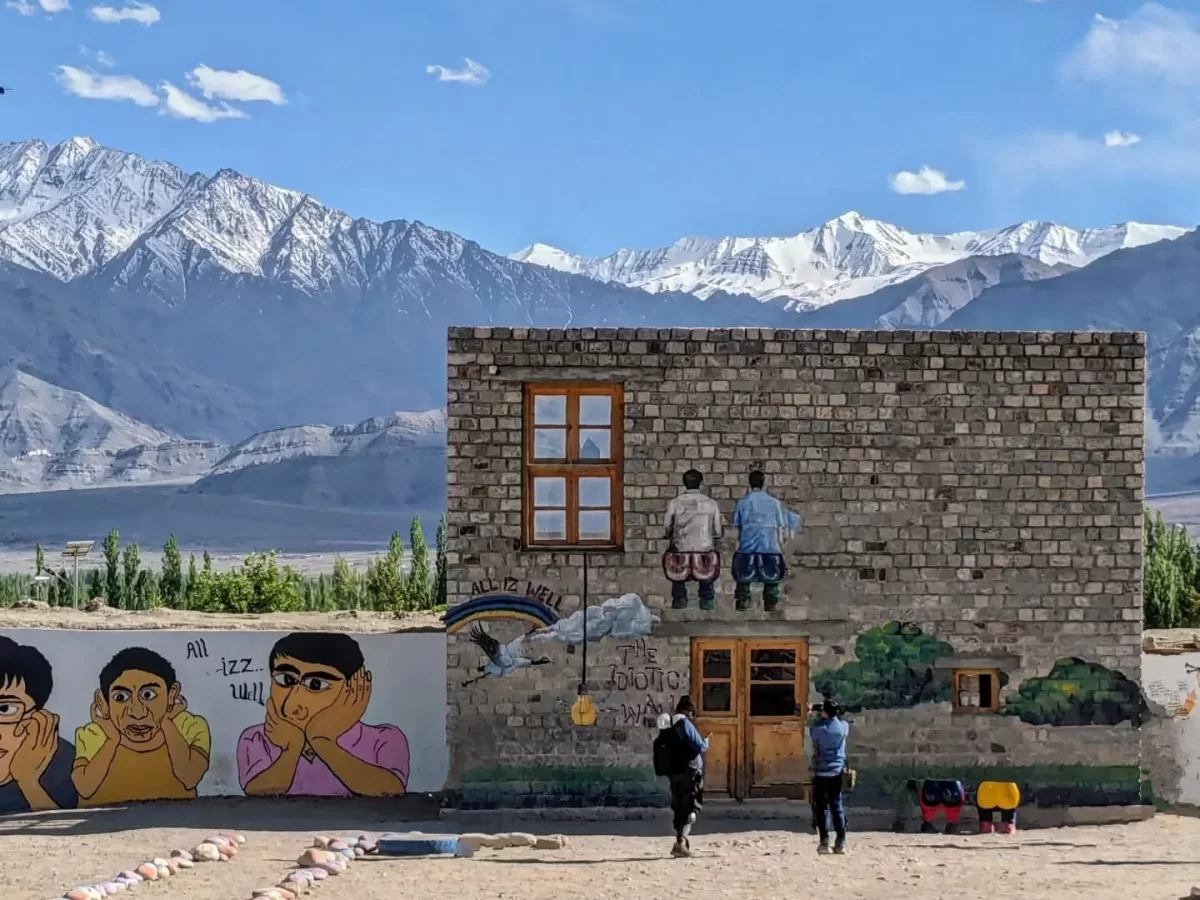 Rancho School mural at Ladakh during clear skies, featuring 3 Idiots wall art snow mountains building visitors, perfect adventure Ladakh tour package.