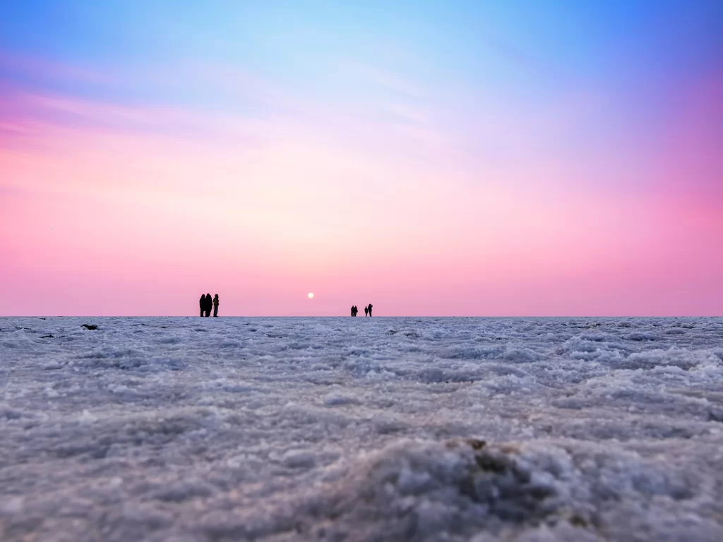 Tourists silhouettes at Rann of Kutch during sunset, featuring pink-blue skies, glowing sun and vast white salt desert, perfect adventure experience with Gujarat tour packages.