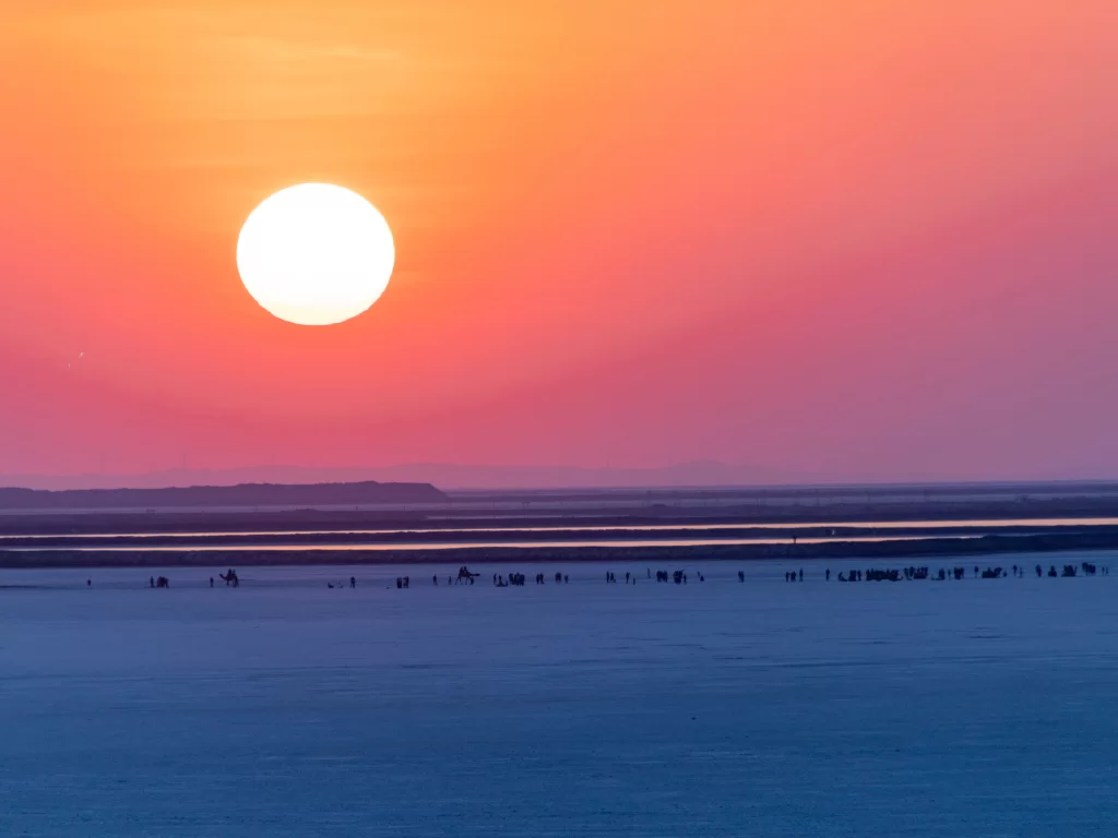 Rann of Kutch sunset during golden hour, featuring silhouetted camels and tourists, orange-pink skies and vast white salt desert, perfect adventure experience with Gujarat tour packages.