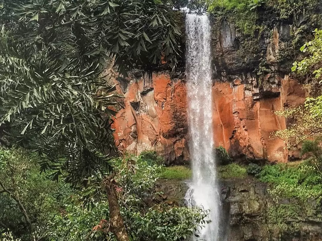 Rautwadi Waterfall in Maharashtra featuring a tall cascading stream flowing down red rocky cliffs surrounded by dense greenery, a scenic monsoon attraction included in Maharashtra tour packages