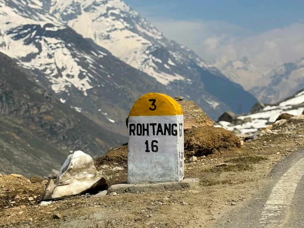 Rohtang Pass milestone sign at Manali Himachal Pradesh during partly cloudy skies, featuring snow peaks and mountain road, perfect adventure experience Himachal Pradesh tour package.