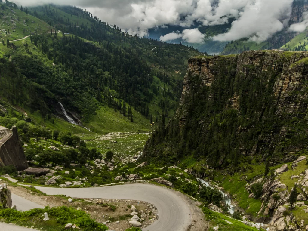 Scenic drive at Rohtang Pass Manali during cloudy weather, featuring winding road waterfalls and lush green valleys with cliffs, perfect adventure experience Himachal Pradesh tour package.
