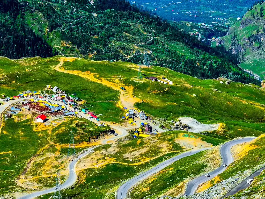 Panoramic view at Rohtang Pass Manali during sunny day, featuring winding roads colorful tents and lush green Himalayan meadows, perfect adventure experience Himachal Pradesh tour package.