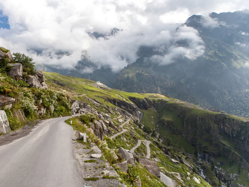 Winding road at Rohtang Pass Manali during misty morning, featuring rugged Himalayan peaks and green meadows, perfect adventure experience Himachal Pradesh tour package.