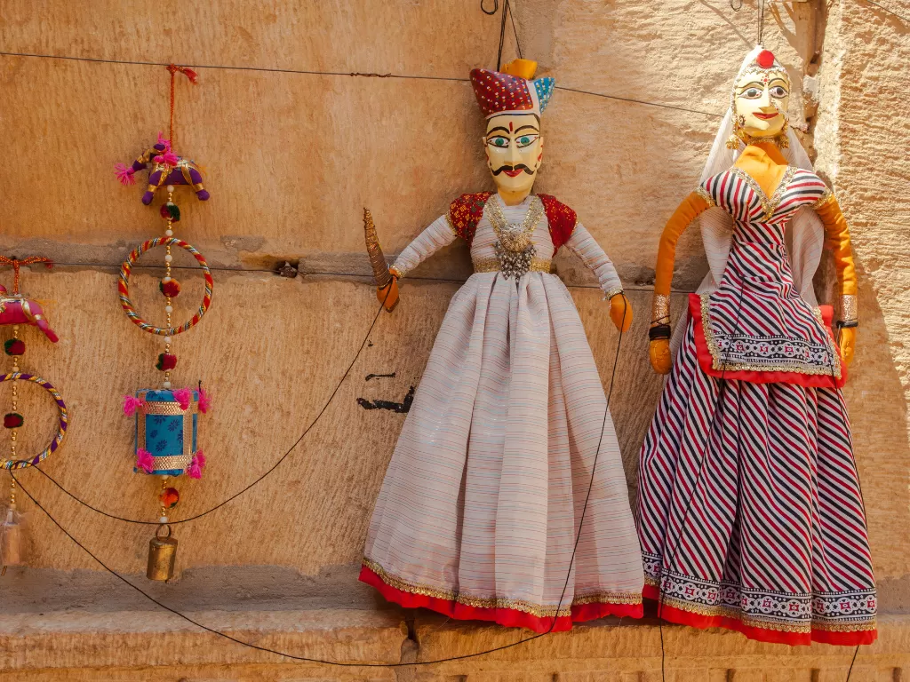 Traditional Rajasthani puppets at Jaisalmer sand dunes during sunset, featuring king queen with turban, sword, skirt on clay wall, perfect cultural experience in Rajasthan tour package.