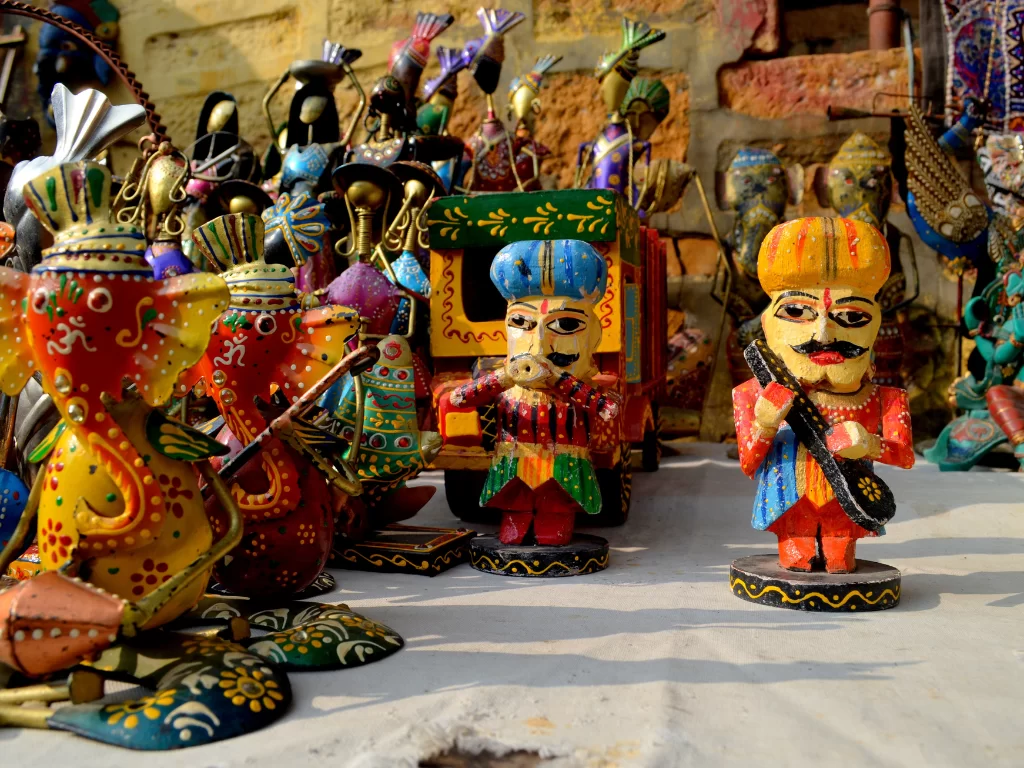 Vibrant Rajasthani folk toys at Jaisalmer sand dunes market, featuring colorful camels, musicians, Ganesha on clay wall backdrop, perfect cultural experience in Rajasthan tour package.