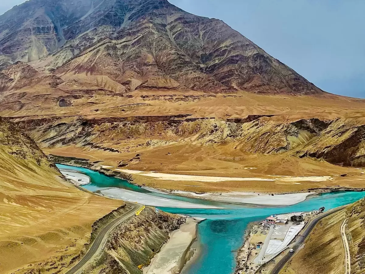 Drone aerial of Indus Zanskar rivers Sangam confluence Leh Ladakh during clear day, featuring turquoise streams merging amid rugged brown mountains, highway, settlements, perfect adventure scenic experience with Sham Valley Leh tour package. 