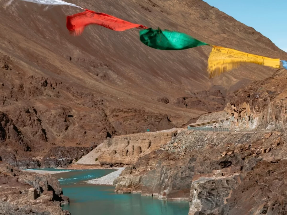 Colorful prayer flags fluttering at Indus Zanskar rivers Sangam Leh Ladakh amid rugged brown mountains, turquoise river, road, perfect spiritual scenic experience with Sham Valley Leh Ladakh tour package.