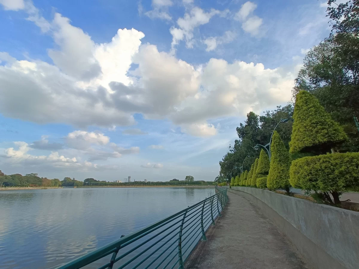 Sankey Tank Bangalore lakeside walkway during sunny day with clouds, featuring green hedges, trees and calm water, perfect Karnataka tour package.