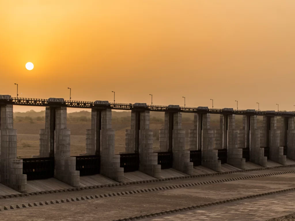 Sant Sarovar Dam at Gandhinagar during sunset, featuring barrage piers silhouetted against orange sky, perfect scenic picnic experience with Gujarat tour packages.