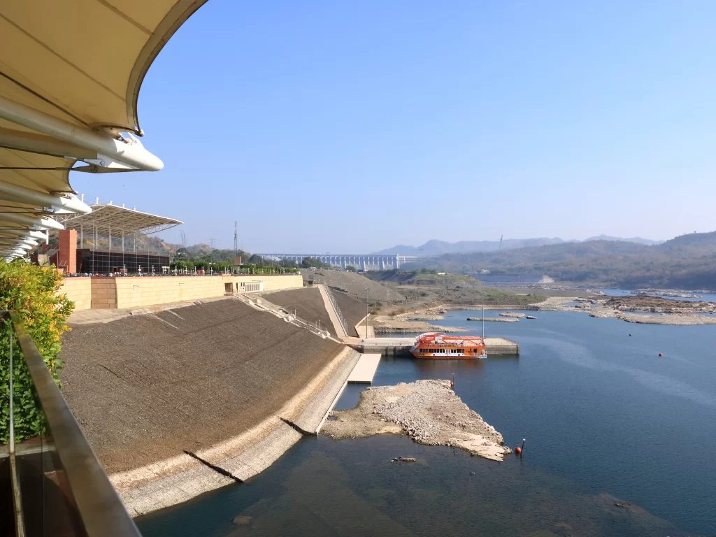 Sardar Sarovar Dam viewing gallery deck during clear day, featuring Narmada reservoir, orange boat, distant dam structure and surrounding hills, perfect engineering marvel with Statue of Unity tour packages.