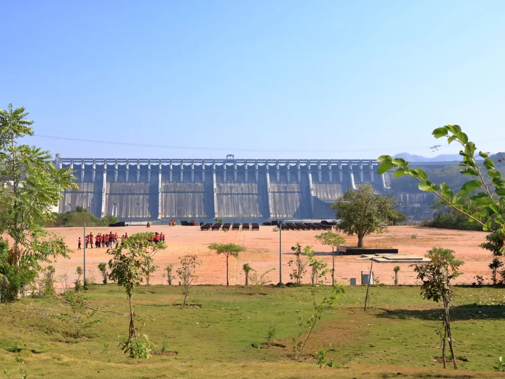 Sardar Sarovar Dam at Kevadia during clear day, featuring massive concrete gravity structure on Narmada River, visitor viewpoint with trees and people, perfect engineering marvel experience with Gujarat tour packages.