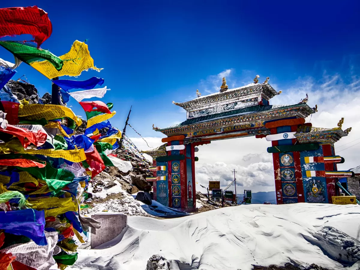 Colorful prayer flags at Sela Pass Tawang during sunny day, featuring Buddhist gate snowy mountains clouds, perfect adventure experience Arunachal Pradesh tour package.