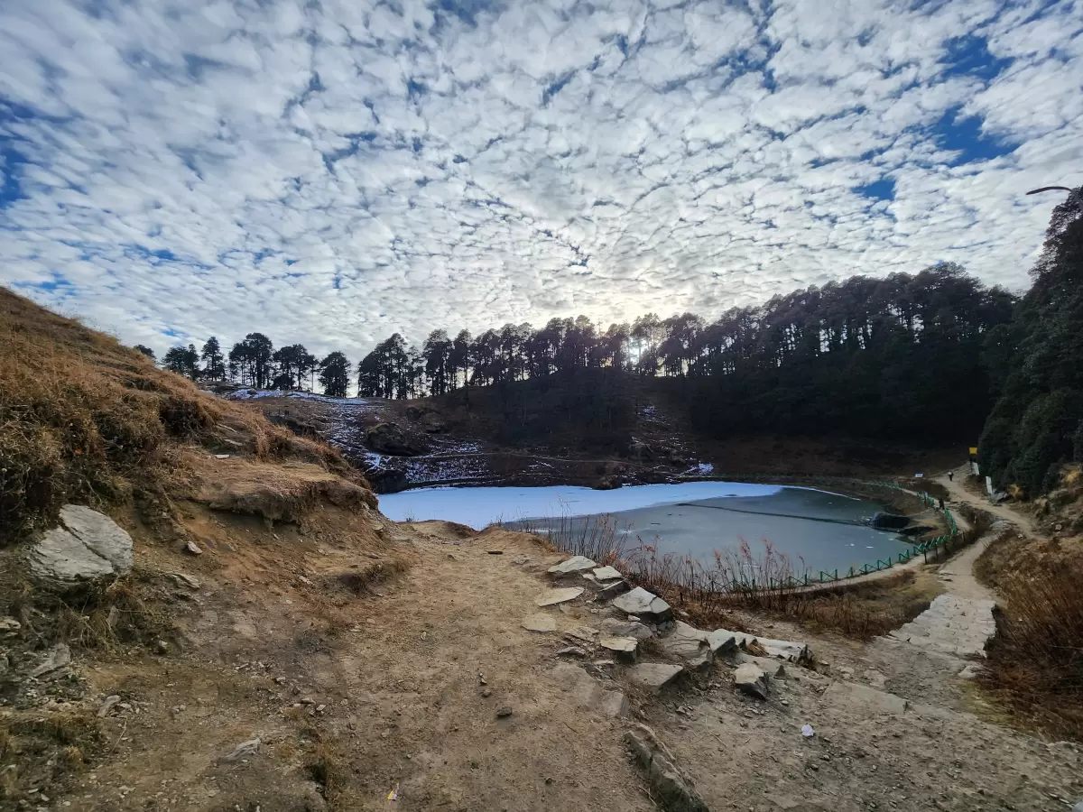 Icy Serolsar Lake Jibhi during winter cloudy skies, featuring pine forests, trek path, rocky shores, perfect serene Himachal Pradesh tour package.